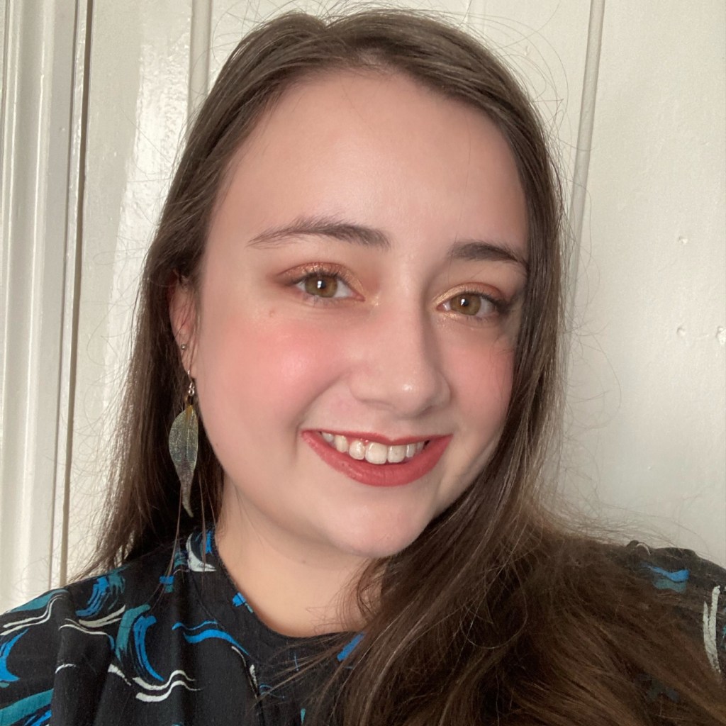 Anna is a smiling white woman with long brown hair, wearing a black dress with blue accents and green drop earrings. She is in front of an off-white background.