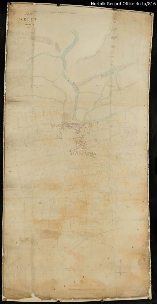 Beige map of Wells-next-the-sea, showing a concentration of buildings in the centre, next to waterways that take up the top third of the map (coloured in blue). The remainder of the map shows roads and large blank rectangular plots, showing how the farmland and pasture of the was area divided up. There are labels on the plan that are too small to read. 