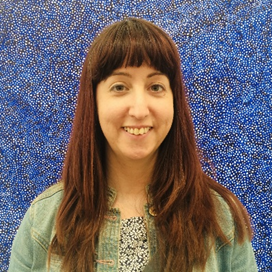 Katy is a smiling white woman with long brown hair, wearing a green jacket in front of a dotty dark blue background.